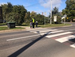 Na zdjęciu widać policjanta umundurowanego z dwójką dzieci przed przejściem dla pieszych - nauka prawidłowego korzystania z przejścia.